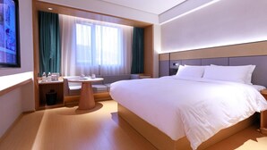 A modern hotel room with a large bed, a small table with a chair, a sofa, and a TV mounted on the wall.