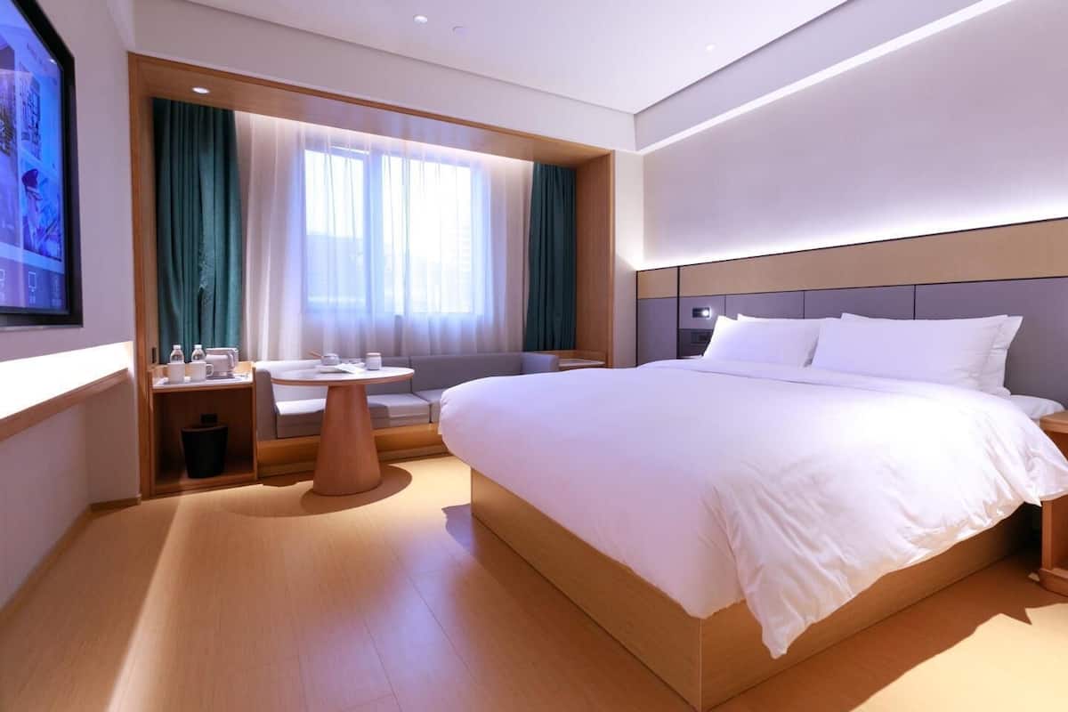 A modern hotel room with a large bed, a small table with a chair, a sofa, and a TV mounted on the wall.