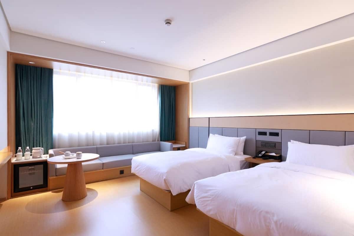 A hotel room with two beds, a small table, a bedside table, a window with curtains, and a wall-mounted control panel.