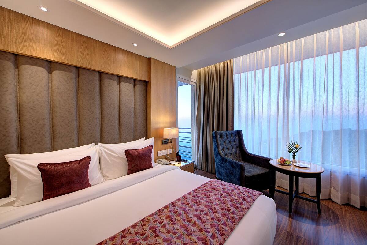 A hotel room with a large bed, a sitting area with a chair and a small table, and a view of the sea through large windows.