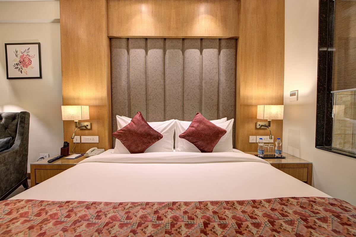 A hotel room with a large bed, two red pillows, a wooden headboard, bedside lamps, a nightstand with a phone and remote controls, and a floral artwork on the wall.