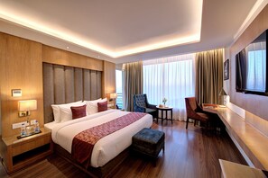 A hotel room with a large bed, bedside tables, a desk, a chair, a TV, and a window with curtains.