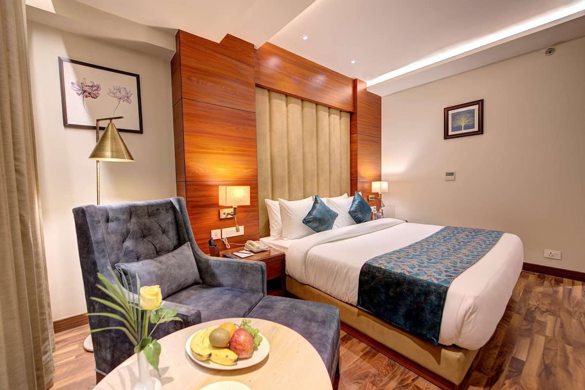 A hotel room with a bed, a chair, a table with fruit, and a lamp.