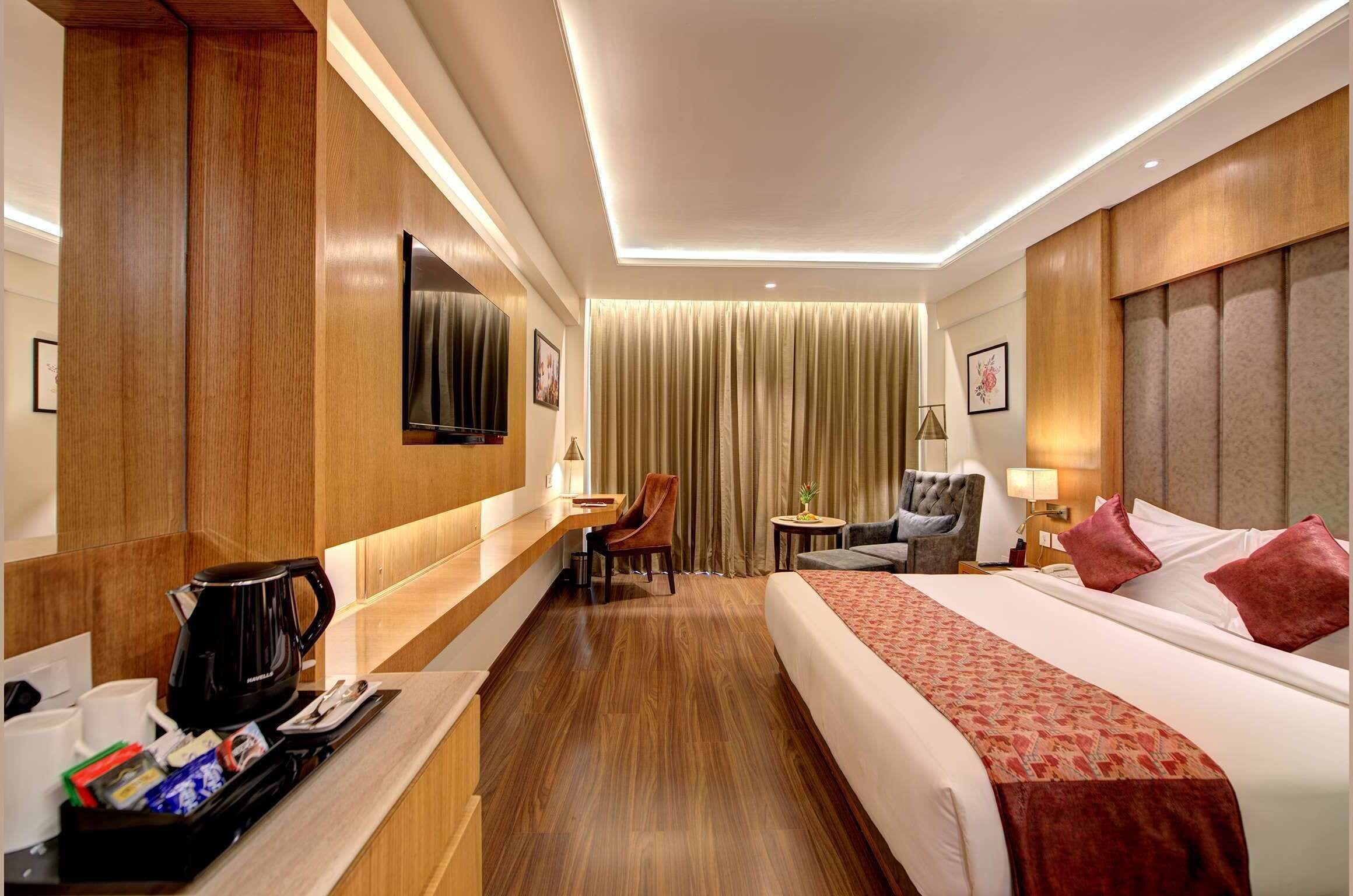 Habitación de hotel con piso de madera, cama con ropa de cama roja, escritorio con cafetera, televisor y pequeña mesa con lámpara.