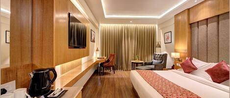 Habitación de hotel con piso de madera, cama con ropa de cama roja, escritorio con cafetera, televisor y pequeña mesa con lámpara.