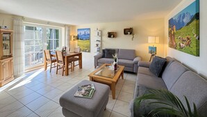 Ein Wohnzimmer mit grauem Sofa, einem Holztisch und einem Essbereich mit Tisch und Stühlen. An den Wänden befinden sich Bilder und im Raum steht eine Pflanze.