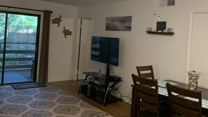 A living room with a ceiling fan, a flat-screen TV on a stand, a dining table with chairs, and a patterned rug. - 2+2 Beach Condo (Fernandina Beach)