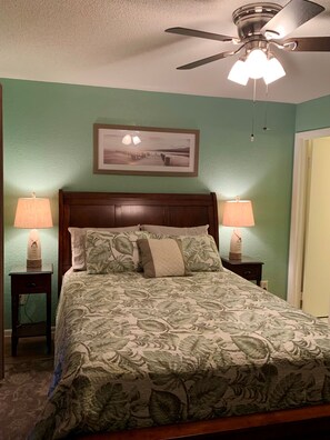 A neatly made bed with floral bedding, two bedside lamps, a ceiling fan, and a framed picture on the wall.