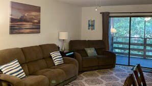 A living room with two brown sofas, a ceiling fan, a painting, a lamp, and a view of greenery through a large window. - 2+2 Beach Condo (Fernandina Beach)