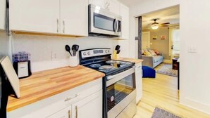 Une cuisine compacte avec des meubles blancs, un four à micro-ondes et une cuisinière. On y trouve un plan de travail en bois et une petite poubelle. La cuisine donne sur un espace de vie avec un canapé bleu et un ventilateur de plafond.
