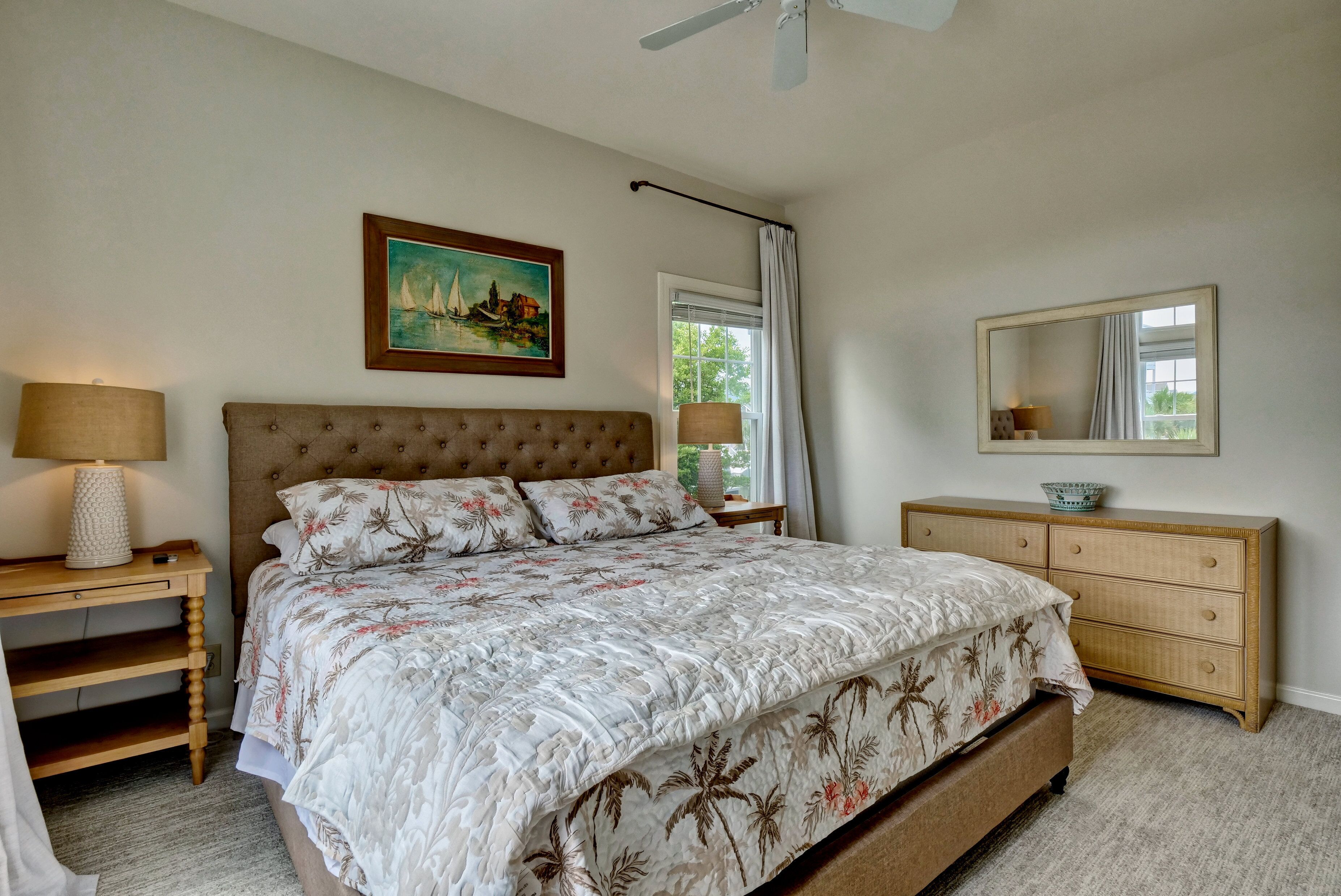 A bedroom with a large bed, a nightstand with a lamp, a dresser, a painting, and a window with curtains.