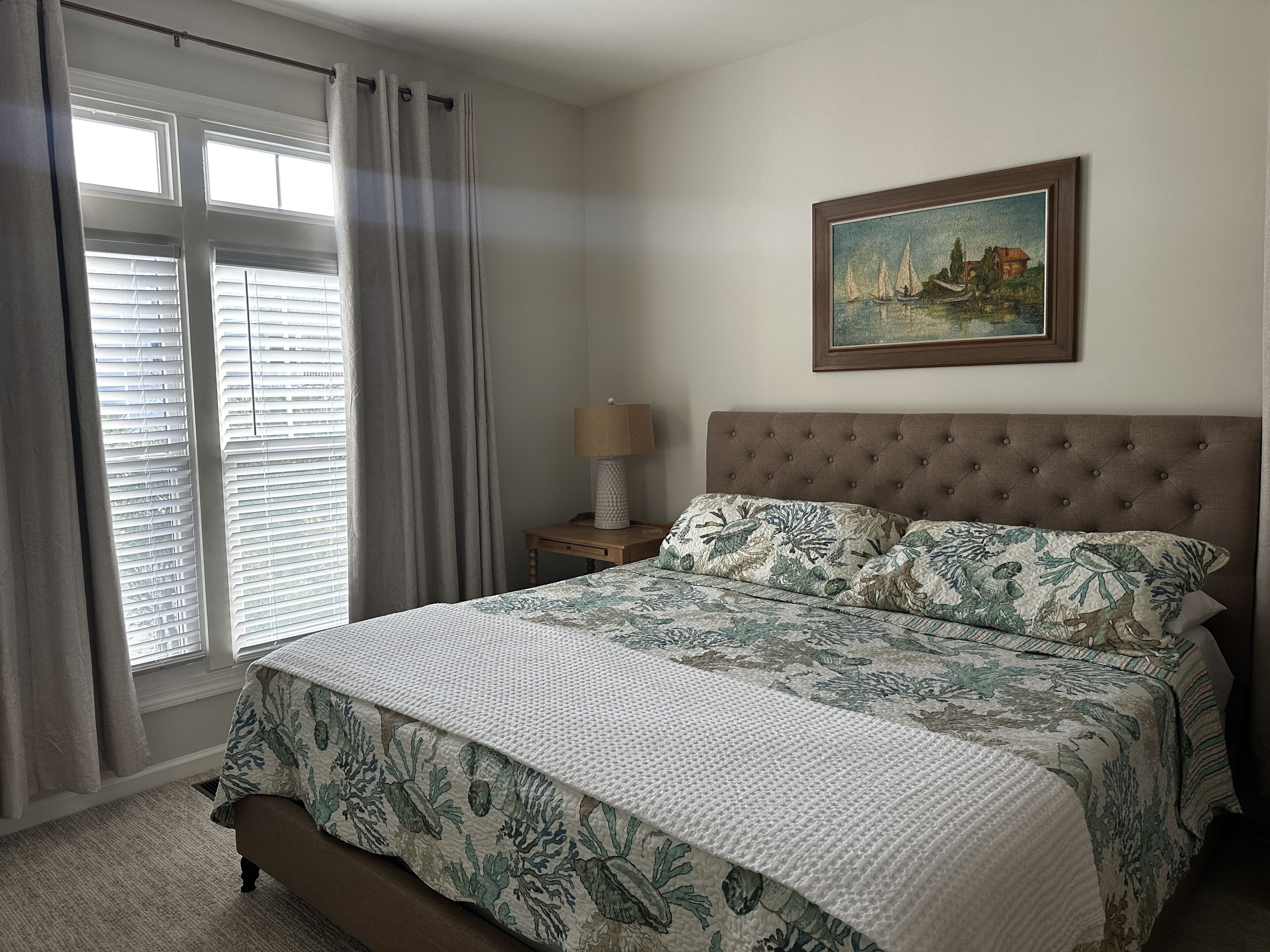 A bedroom with a large bed, a nightstand with a lamp, a window with blinds, and a painting on the wall.
