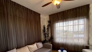 Una sala de estar con sofá, una mesa de centro, una silla, un ventilador de techo y un ventanal con cortinas.