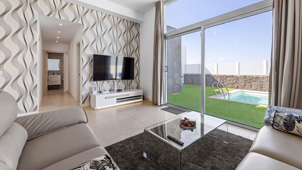 A modern living room with a flat-screen TV, a glass coffee table, and a view of a swimming pool and garden. - Villa La Sombrera (Playa Blanca)