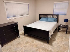 A bedroom with a bed, dresser, nightstand, lamp, and two windows with blinds.