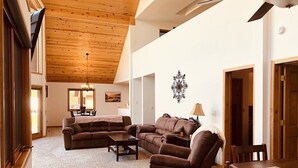 A spacious living room with a high ceiling and wooden beams, furnished with a brown sofa set, a coffee table, and a dining table with chairs.