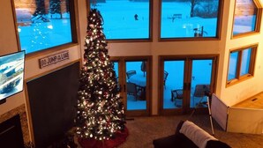 A room with a large Christmas tree, a fireplace, and multiple windows overlooking a snowy outdoor area.