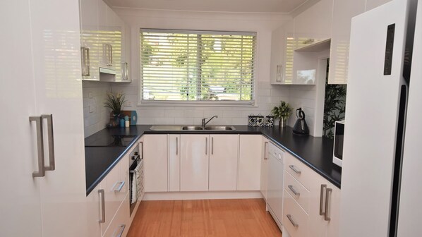 A modern kitchen with white cabinetry, a black countertop, and a window with blinds. - Riverwood - Beautiful destination, large home perfect for a family getaway! (Nambucca Heads)