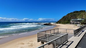 A beach with a wooden walkway leading to a building with a green roof.