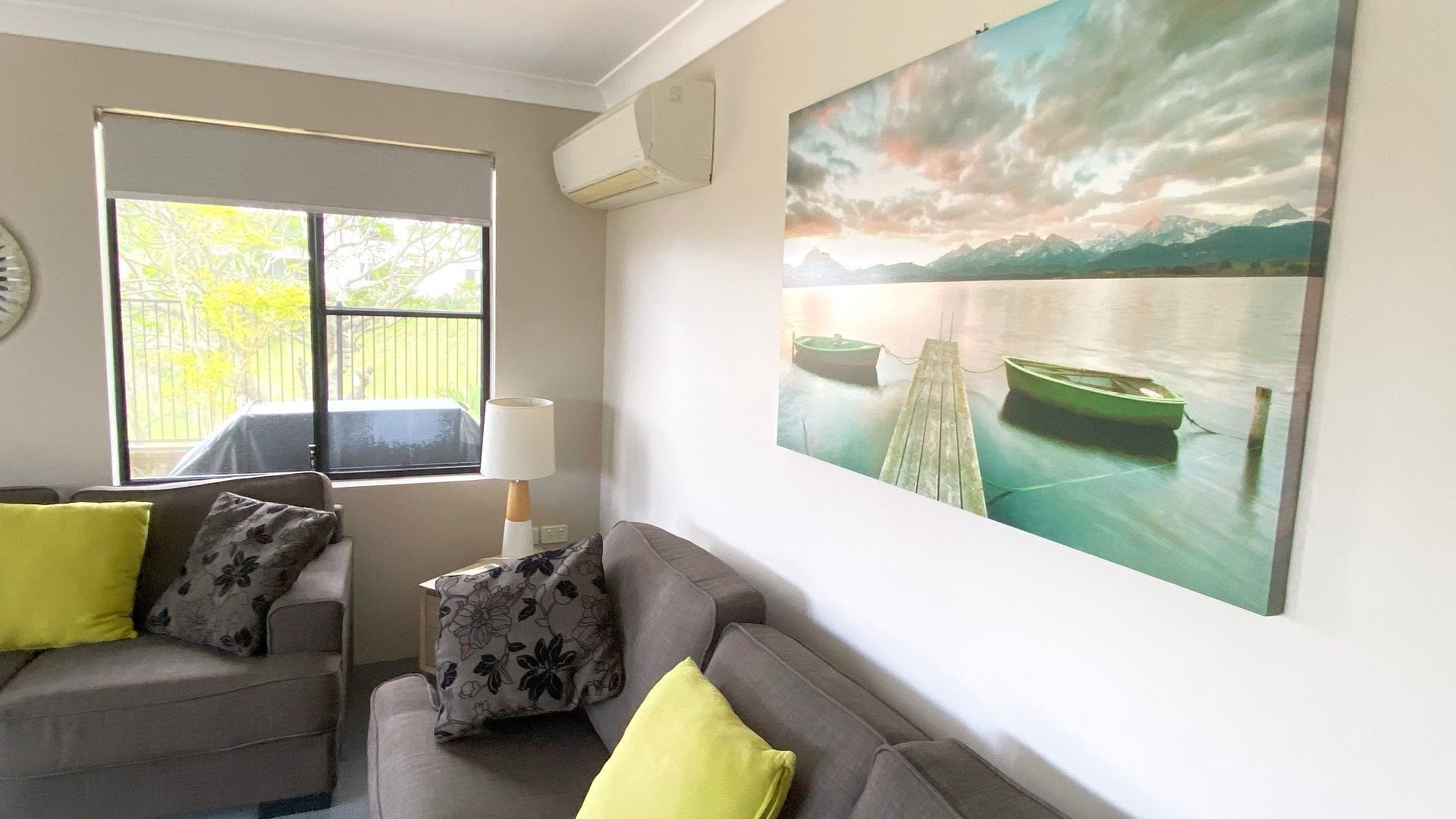 A living room with a grey sofa, two yellow cushions, a lamp, and a large landscape painting of a lake and mountains.