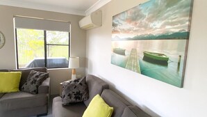 En stue med en grå sofa, to gule puder, en lampe og et stort landskabsmaleri af en sø og bjerge.