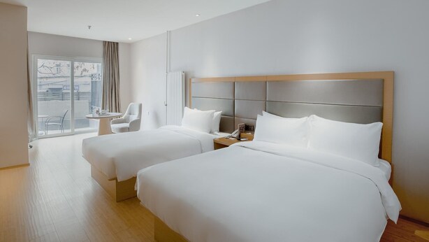 A hotel room with two beds, a small table, a chair, and a window with curtains.