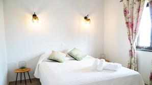 Un lit bien fait, avec une literie blanche et deux oreillers, une petite table d’appoint sur laquelle se trouvent un récipient en verre et une plante, et deux appliques murales.