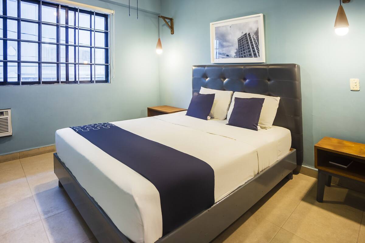 Una cama bien hecha con un cabecero oscuro, una ventana con persianas, un cuadro en la pared y una mesita de noche pequeña.