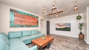 A living room with a teal sofa, a wooden coffee table with wheels, a rustic wooden wall, a green hanging light fixture, and a landscape painting.