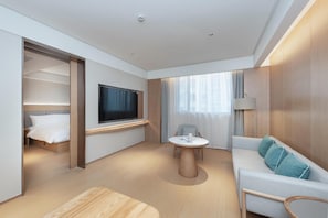 A modern hotel room with a bed, a TV, a sofa, a small table, and a window with curtains.