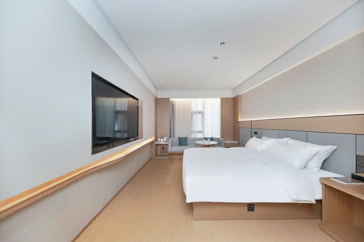 Une chambre d’hôtel avec un grand lit, un téléviseur fixé au mur, une table de chevet, une petite table avec une chaise et une fenêtre avec des rideaux.