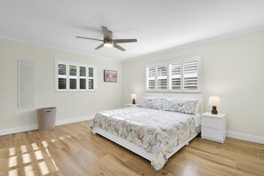 Un dormitorio con cama, dos mesitas de noche con lámparas, un ventilador de techo, una ventana con persianas y un cubo de basura.