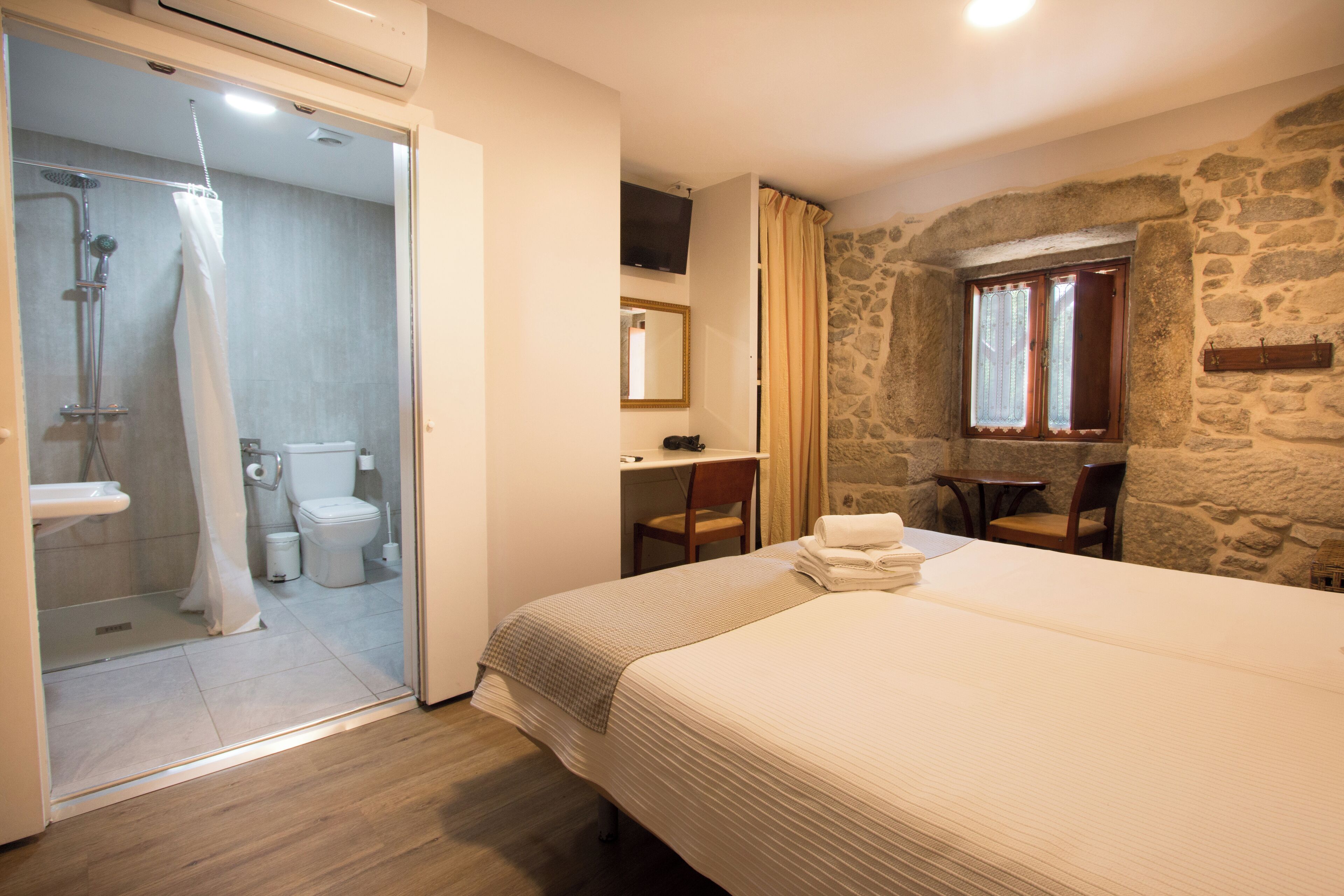 A hotel room with a bed, a desk, a chair, a stone wall, a window, and a bathroom with a shower and toilet.