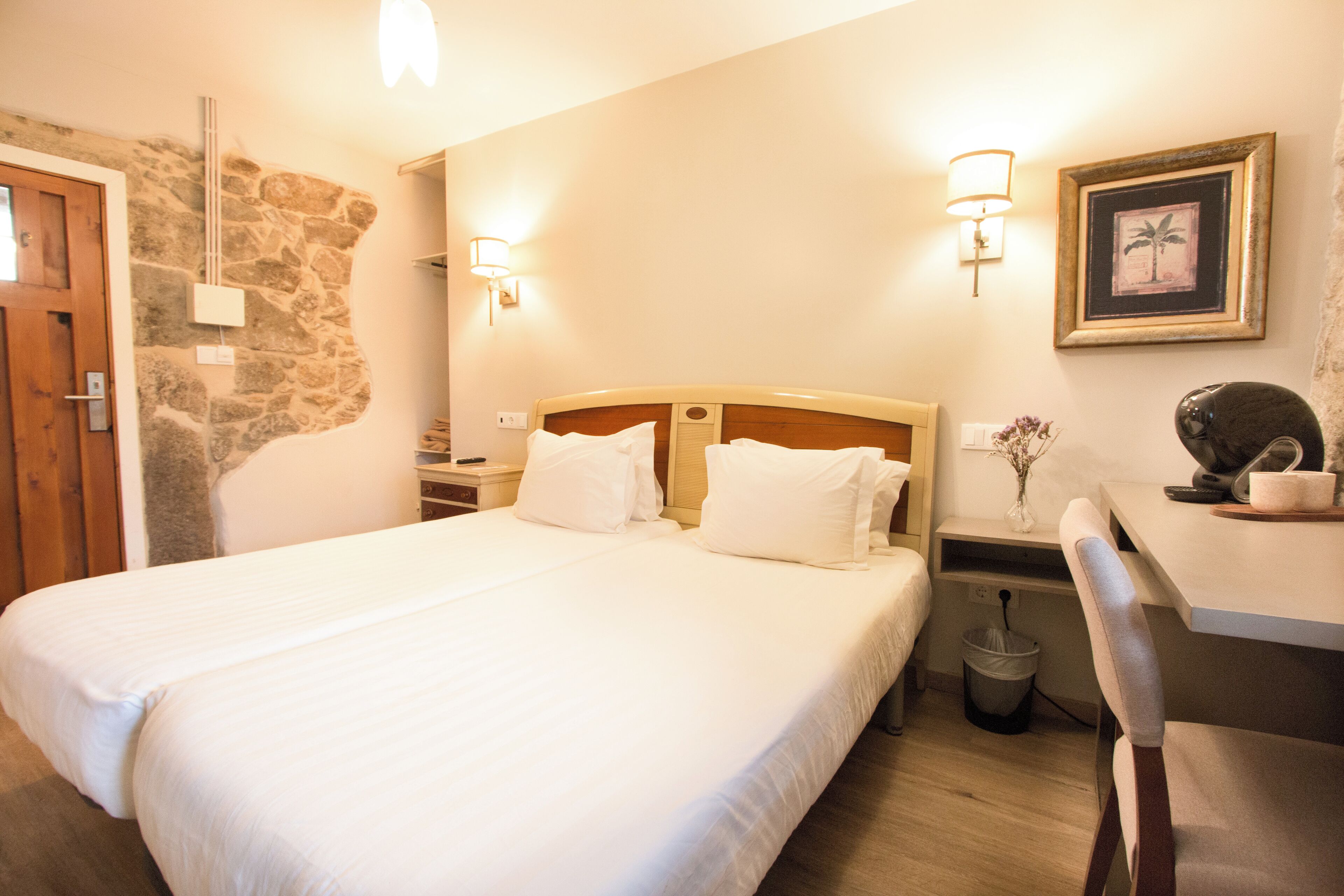 A hotel room with a large bed, a wooden headboard, two bedside tables with lamps, a small desk with a chair, a trash bin, and a framed picture on the wall.