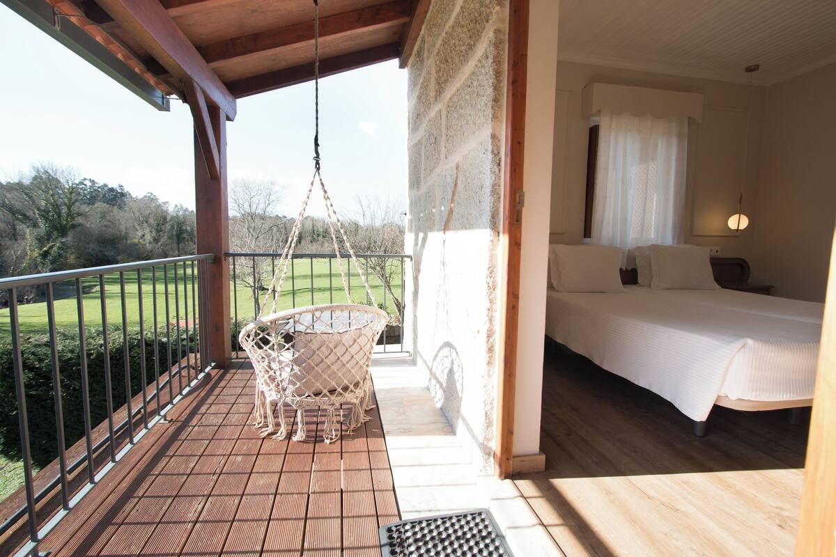 A balcony with a hanging chair, a bed with white bedding, and a view of a green landscape.