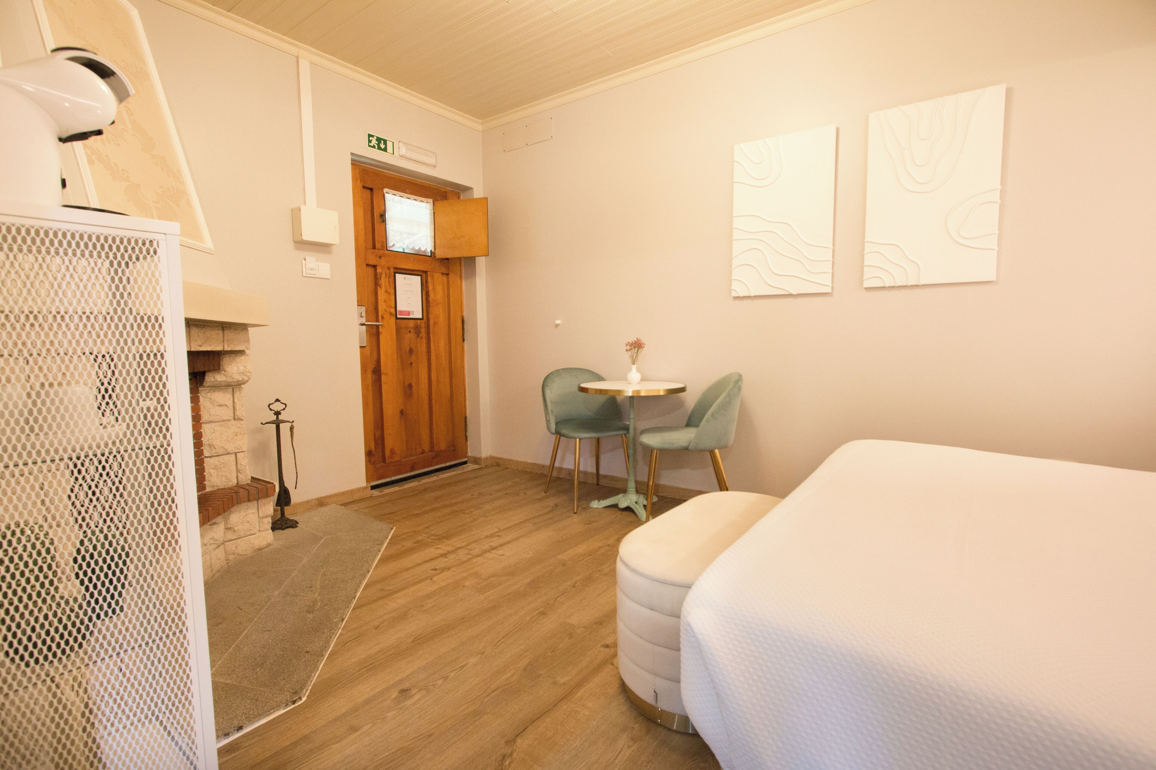 Una habitación de hotel con piso de madera, una cama, una pequeña mesa redonda con dos sillas y una puerta con un cartel encima.