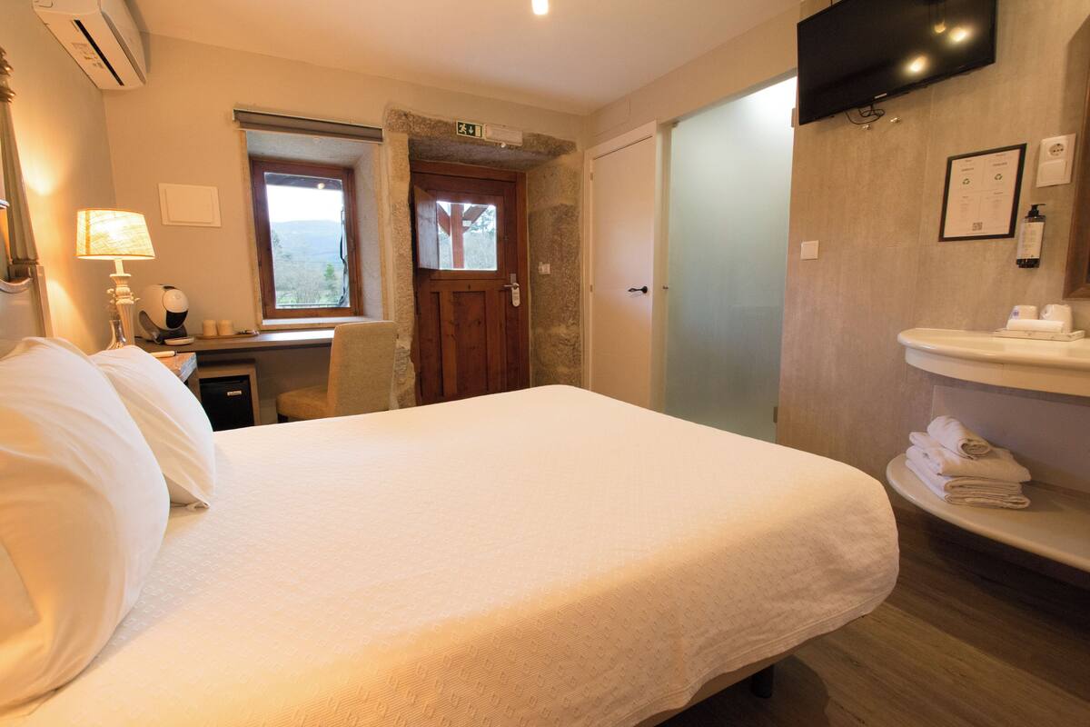 A hotel room with a large bed, a desk with a lamp, a TV mounted on the wall, and a bathroom with a sink and towels.