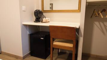 Quarto compacto com uma pequena geladeira preta, uma cadeira de madeira, uma bancada branca, uma cafeteira e um espelho com moldura dourada.