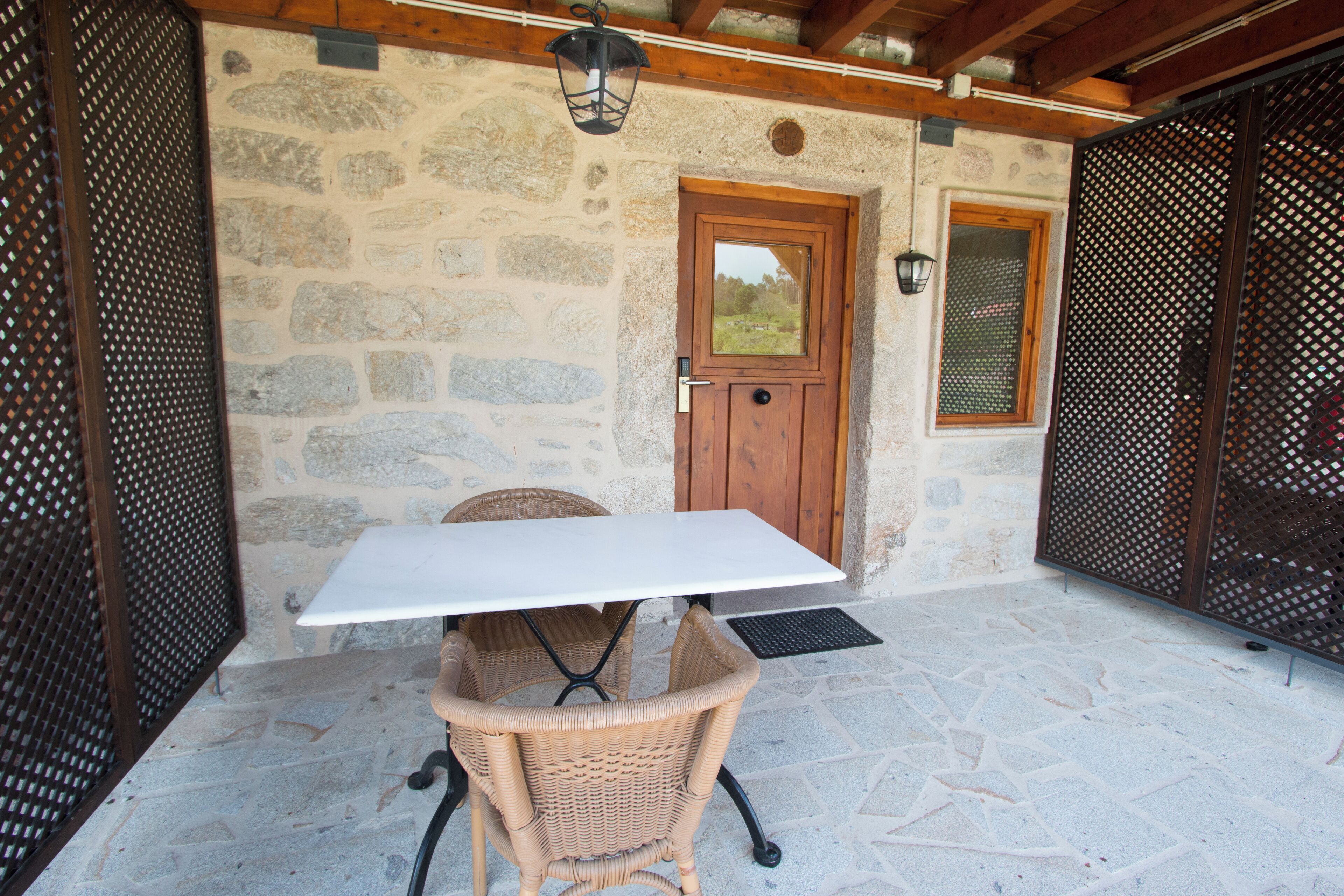 A stone-walled outdoor patio with a table and wicker chairs, a wooden door, and a window with a view of the countryside.