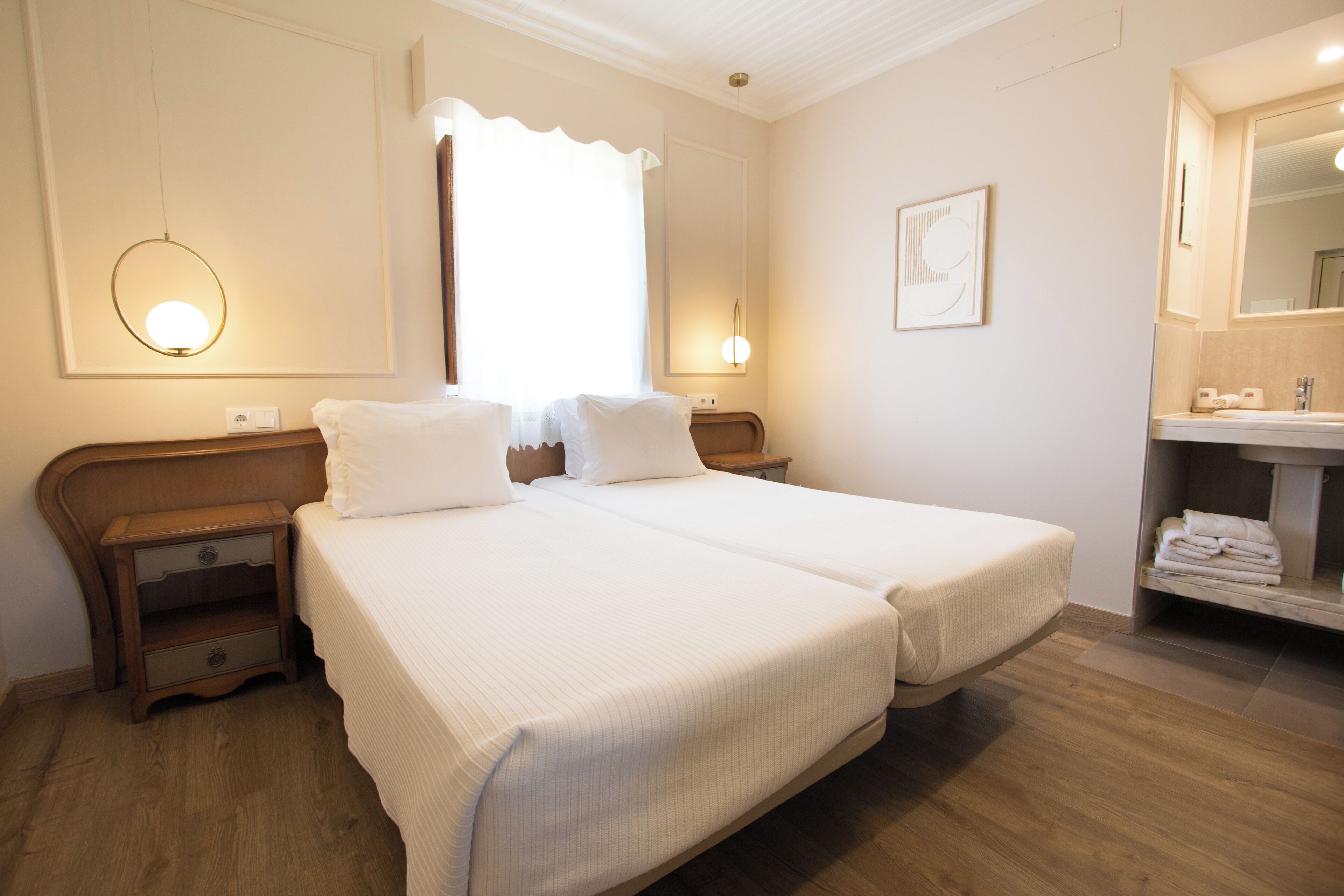 A hotel room with two beds, a nightstand, a mirror, and a bathroom with a sink and towel rack.