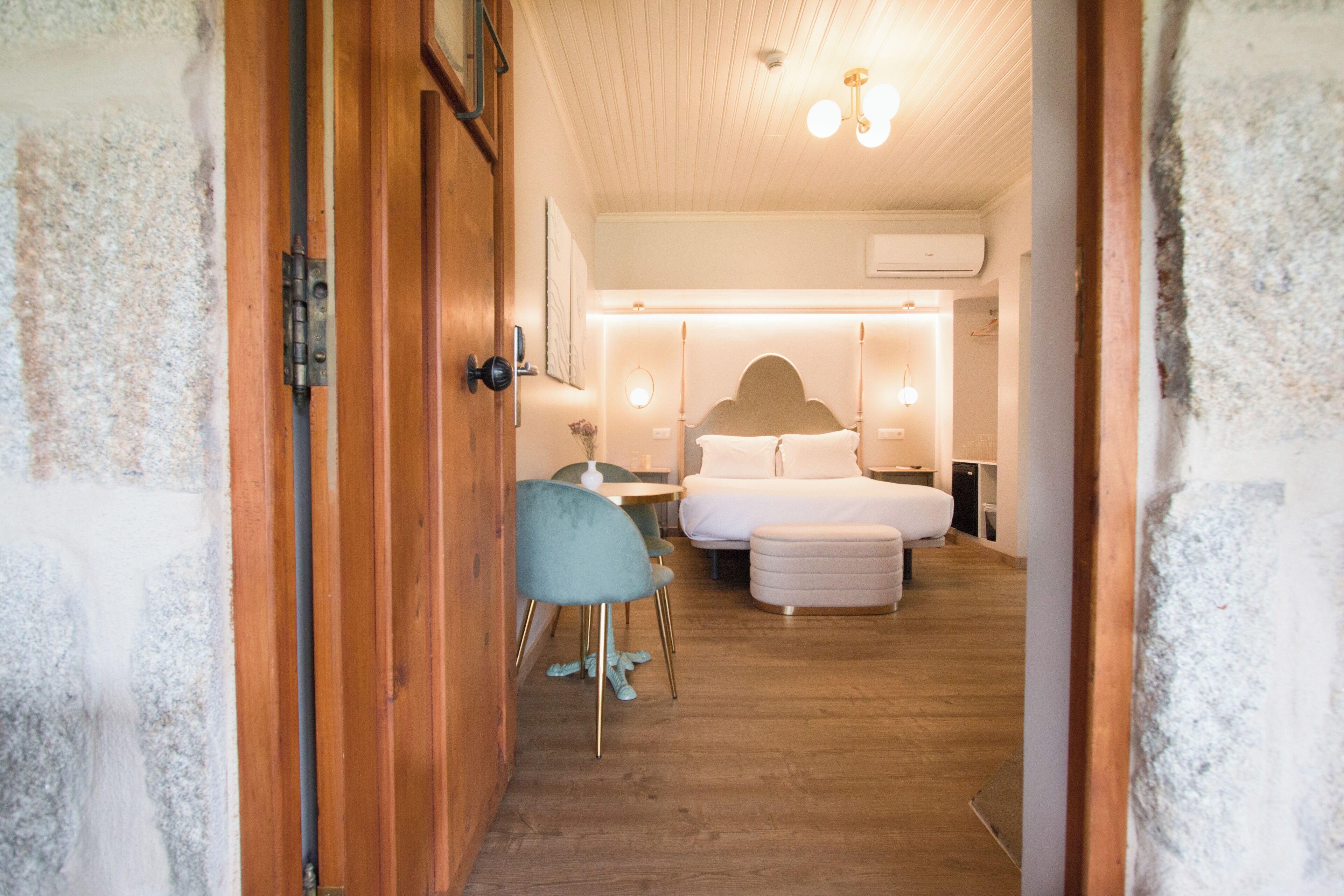 A hotel room with a wooden door, a bed with white bedding, a small table, a chair, and a bedside table with a lamp.