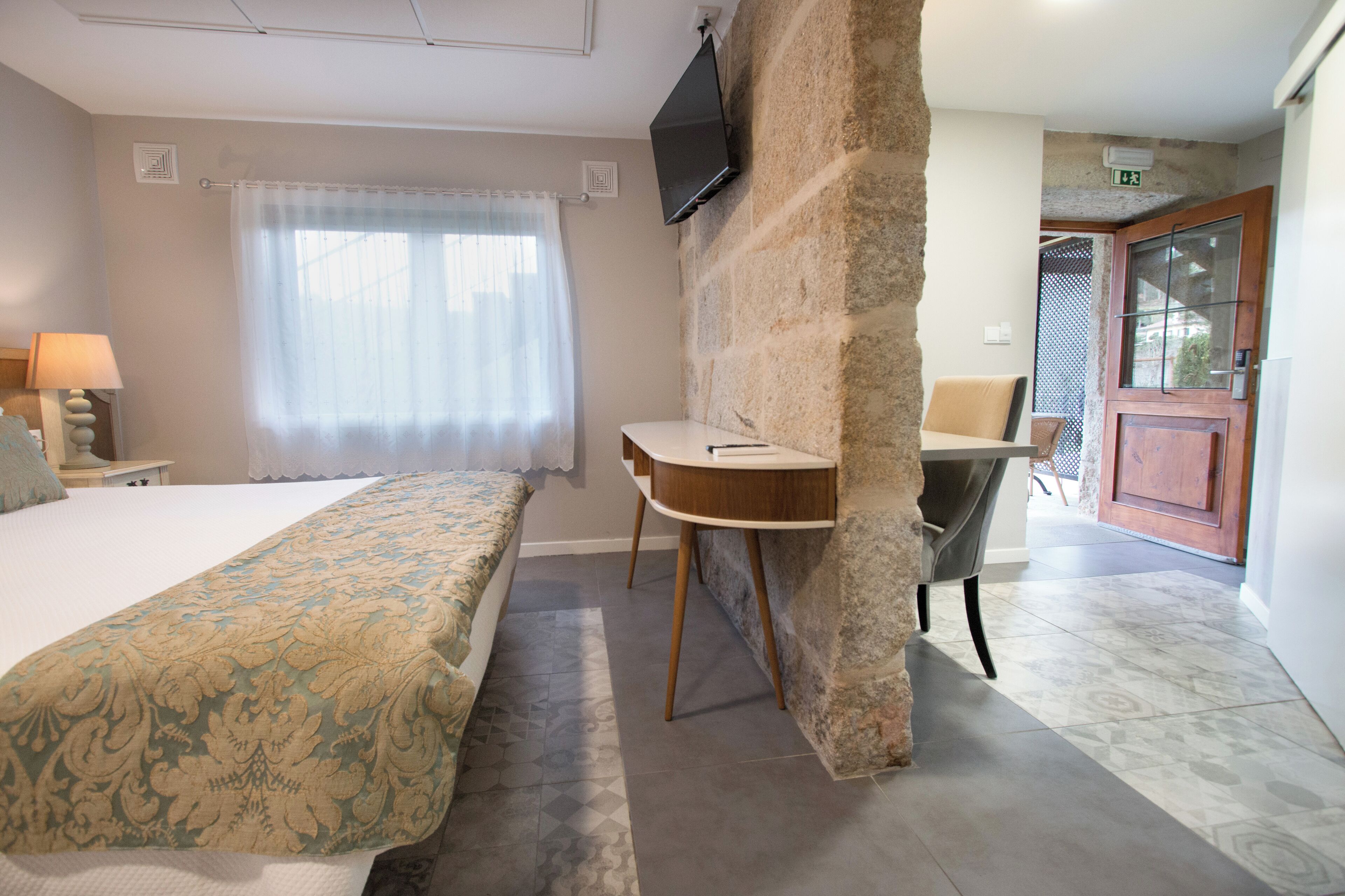 A hotel room with a bed, a desk, a chair, a television mounted on a stone wall, and a view of the outside.