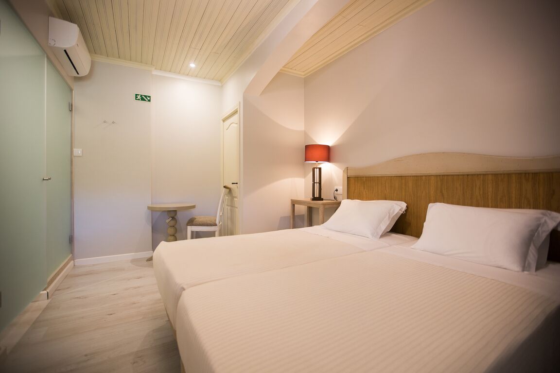 Habitación de hotel con una cama grande, una mesita de noche con lámpara, una pequeña mesa y una silla. Se observa un equipo de aire acondicionado en la pared y una puerta con señal de salida.