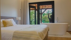 Un dormitorio con cama, bañera y balcón con vistas a la vegetación.