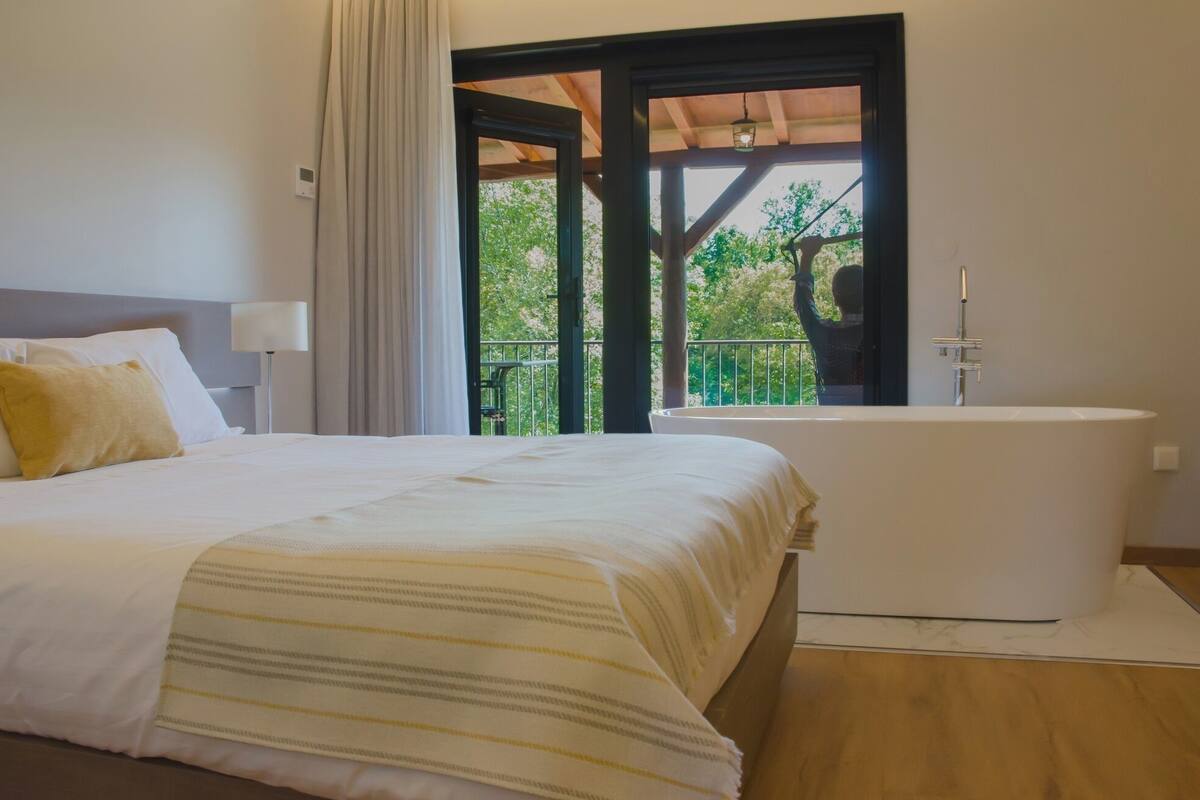 A bedroom with a bed, a bathtub, and a balcony with a view of greenery.