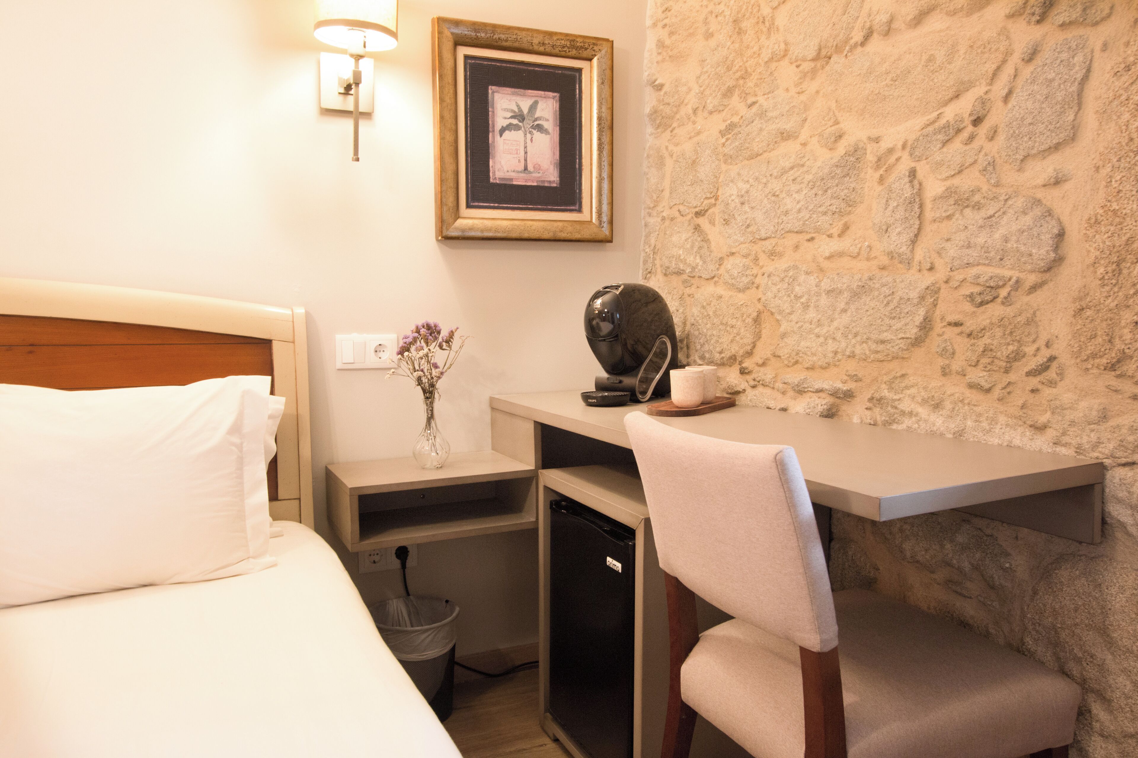Una camera d'albergo compatta con un letto, una scrivania, una sedia, un cestino e una parete in pietra.