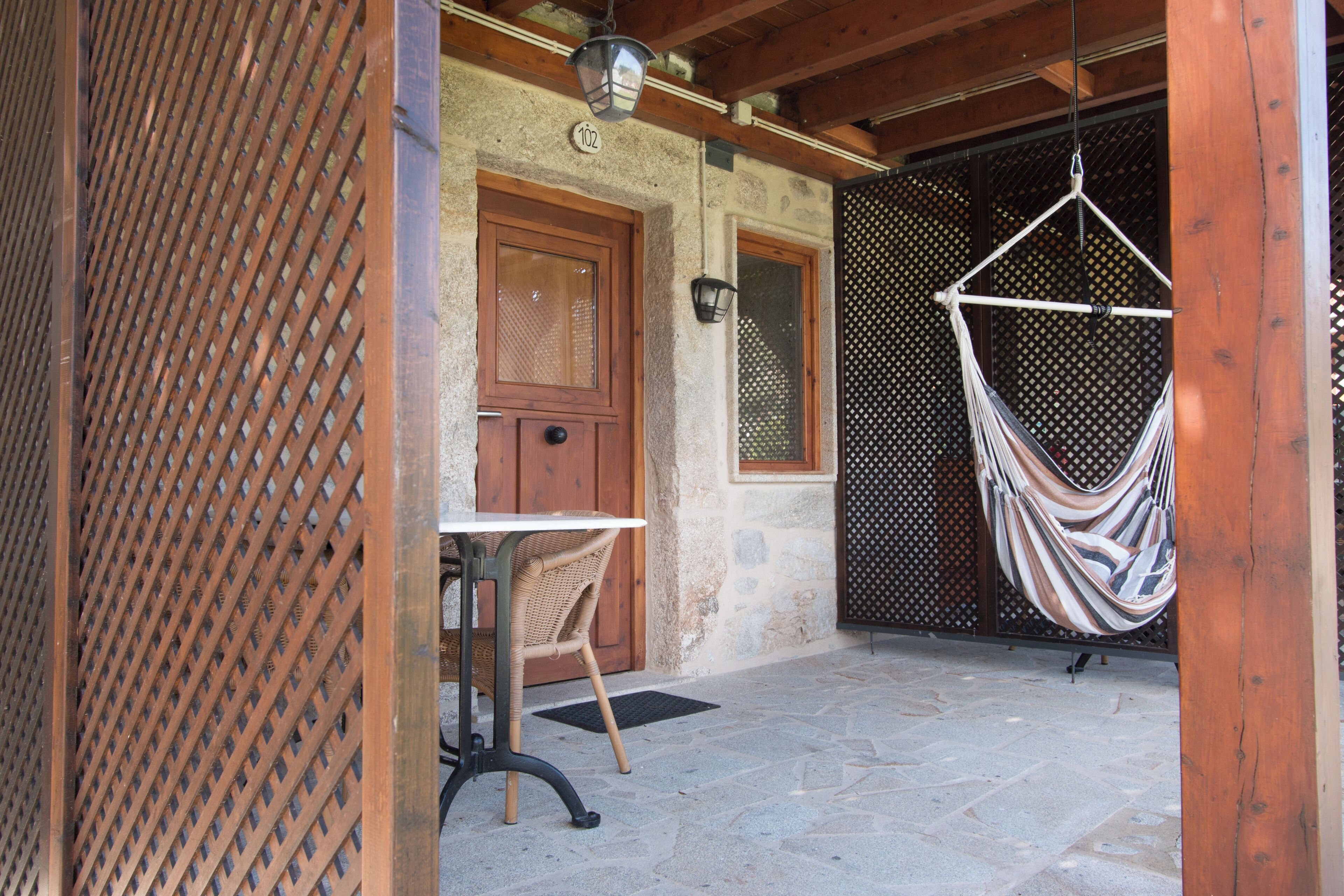 A stone building with a wooden door, a hanging hammock, and a wicker chair outside.