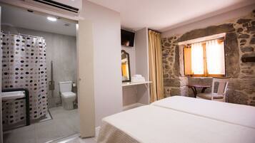 A hotel room with a bed, a bathroom with a shower curtain, a toilet, and a stone wall with a window.