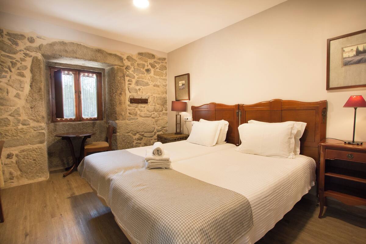 A bedroom with a stone wall, a wooden bed, a small table, and a lamp.