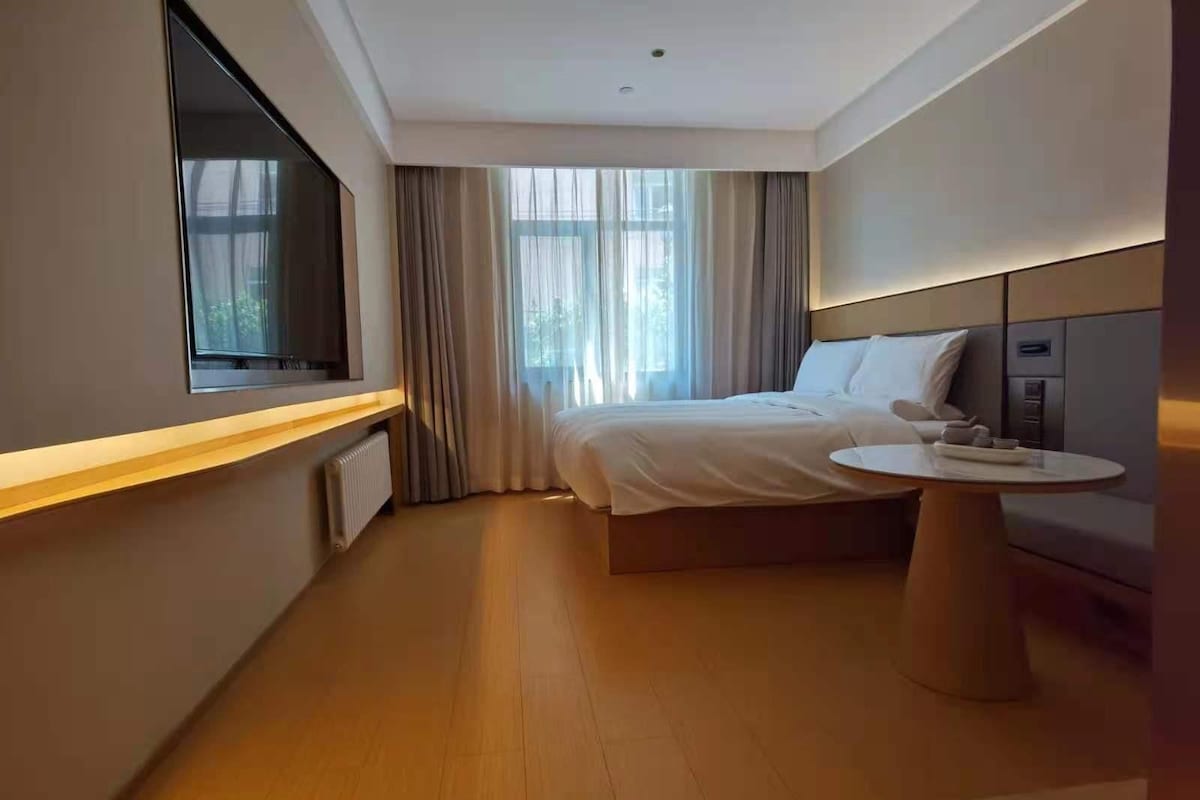 A modern hotel room with a large bed, a bedside table, a TV, and a window with curtains.